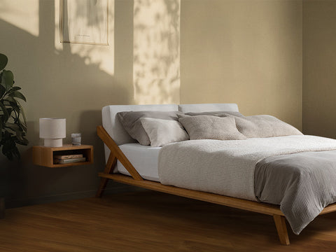nordic space bett und nachttisch aus eiche massivholz mit rückenpolster in farbe natur viele kissen und graue bettdecke auf matratze in schlafzimmer mit sonne
