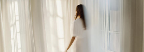 Frau mit weißem Kleid zwischen weißen im Wind wehenden Vorhängen vor Balkontür einer Altbauwohnung